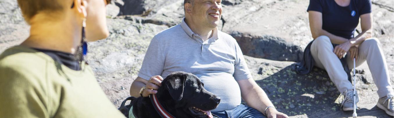 Three smiling people sitting on a rock with a black guide dog.